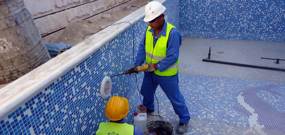 Two workers in safety gear from pool plaster companies repair equipment on the tiled wall of an empty swimming pool.