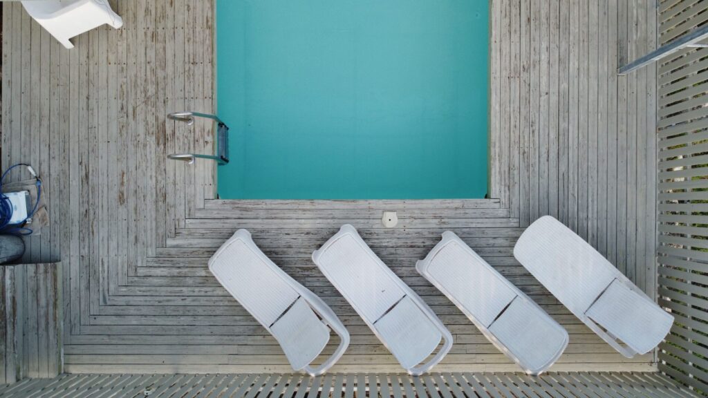 Four white lounge chairs are arranged beside a rectangular swimming pool on a wooden deck, viewed from above.