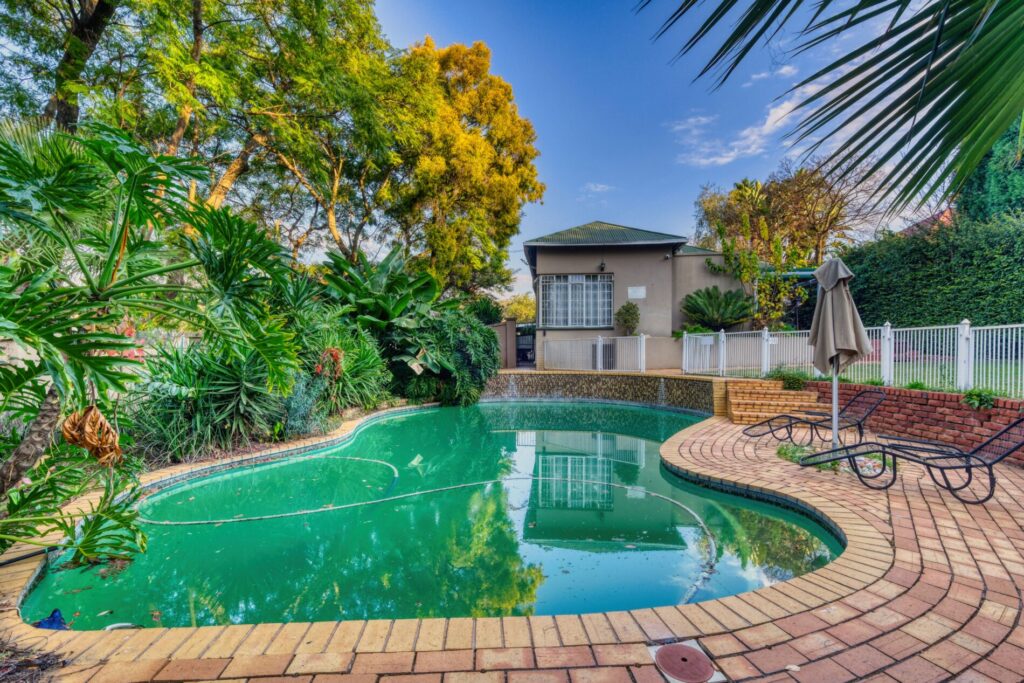Backyard with a kidney-shaped pool, brick patio, lounge chairs, umbrella, and lush greenery surrounding the area.