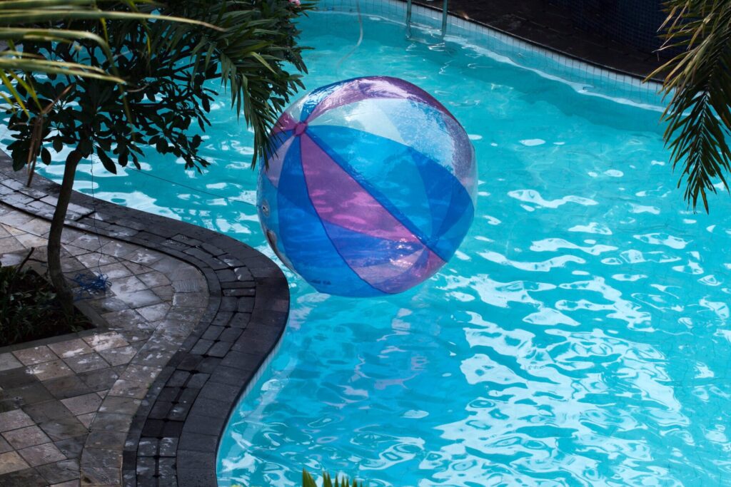A large, colorful beach ball floats in a clear blue swimming pool next to a paved poolside area with plants.