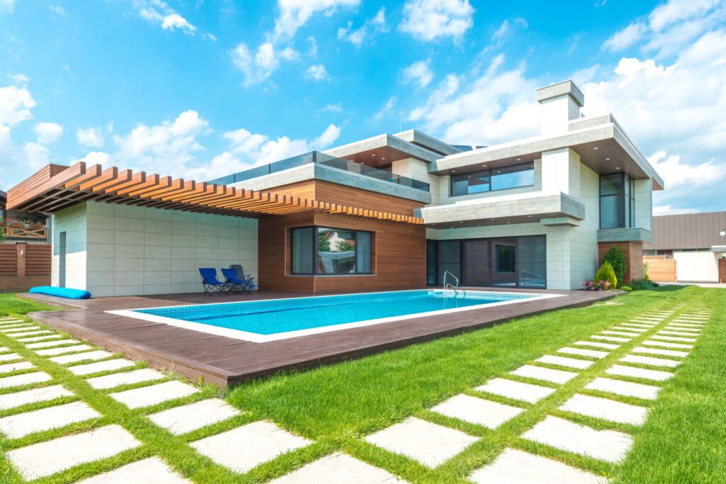 Modern two-story house with a wooden deck, swimming pool, and green lawn under a blue sky with scattered clouds.