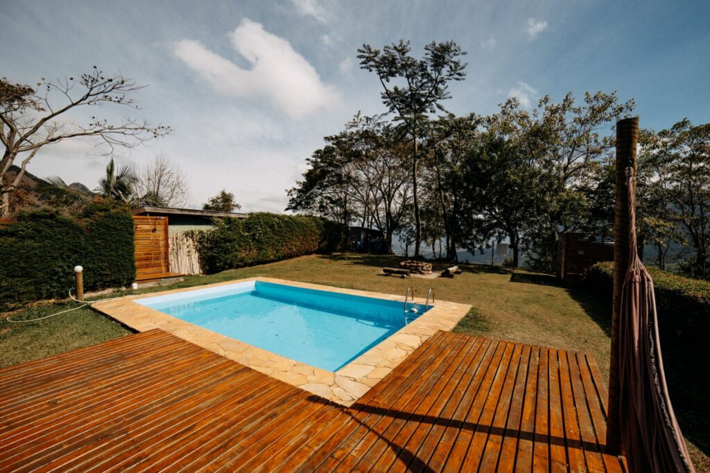 Rectangular swimming pool with stone border, surrounded by grass, trees, wooden deck, and clear blue sky.