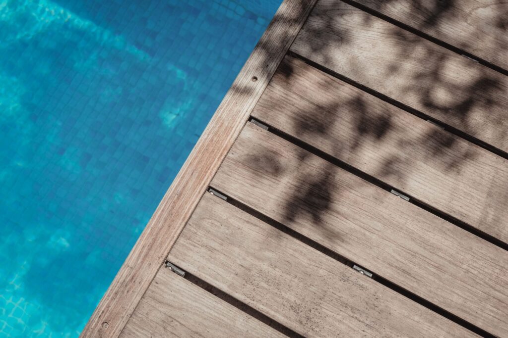 Wooden deck next to a clear blue swimming pool, with shadows of tree branches on the deck.