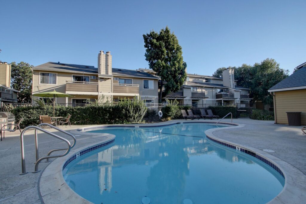 Outdoor swimming pool with poolside chairs in front of two-story apartment buildings on a clear day.