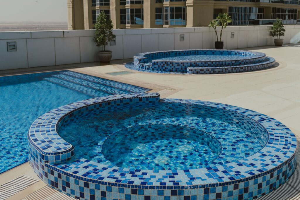 Two circular blue-tiled hot tubs beside a rectangular swimming pool on an outdoor terrace with potted plants.