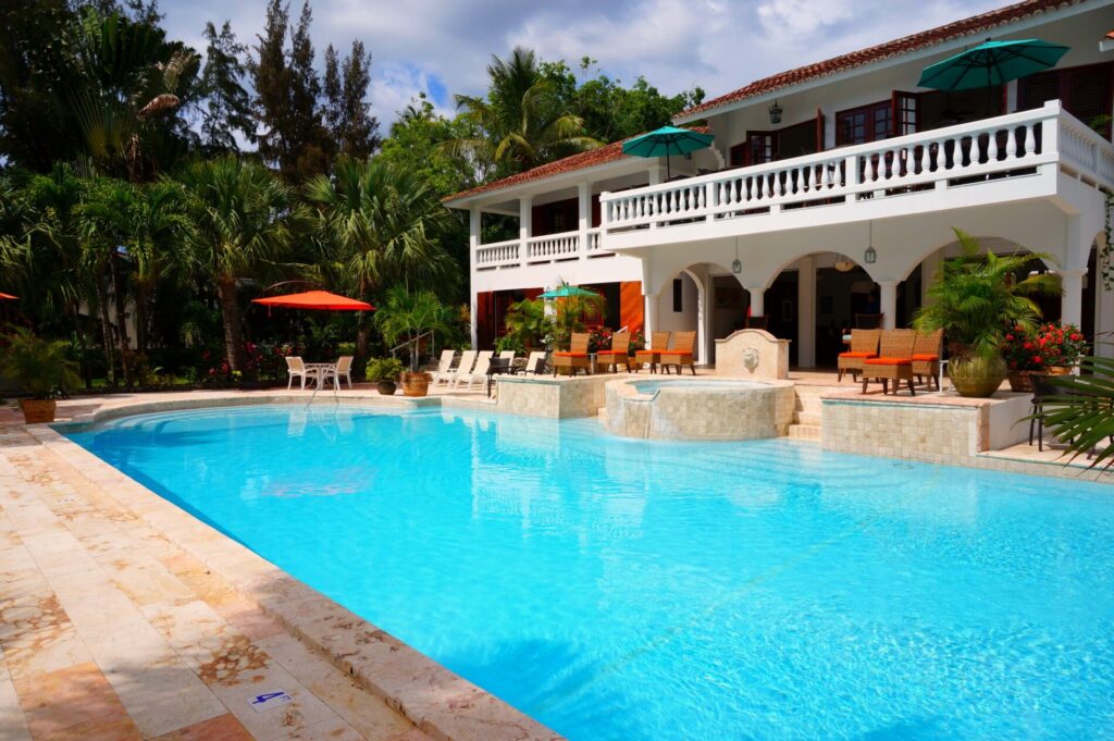 Large outdoor swimming pool with lounge chairs, umbrellas, and a two-story house surrounded by tropical plants.