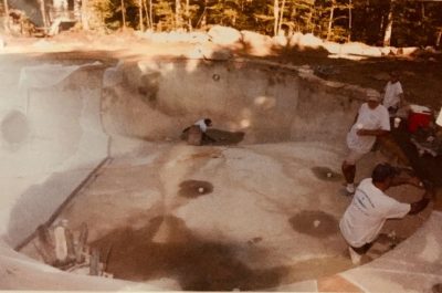 Three workers are resurfacing and repairing an empty, unfinished concrete swimming pool outdoors in daylight.