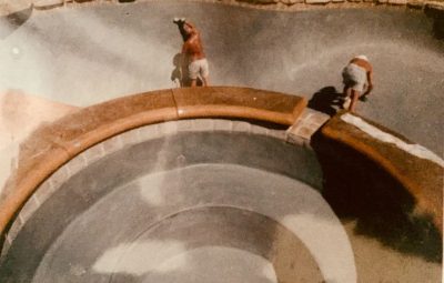 Two workers apply material to the inside surface of an empty, circular swimming pool under construction.