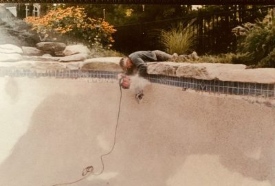A person lies on the edge of an empty pool, using a power tool to work on the pool wall.