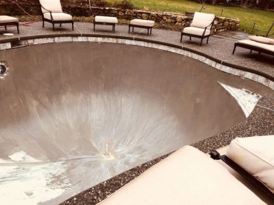 Empty in-ground pool with smooth concrete surface, surrounded by patio chairs on a stone patio in a backyard.