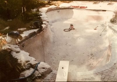 Empty swimming pool under maintenance; two people work on stone edges, tools and equipment scattered inside.