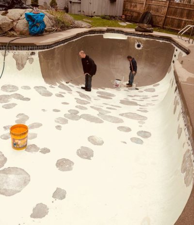 Two people work inside an empty, partially patched swimming pool with tools and a yellow bucket nearby.