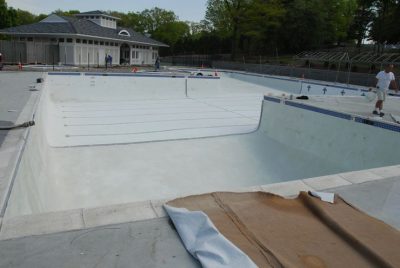 Empty outdoor swimming pool under construction, with workers and unfinished surfaces visible around the pool area.