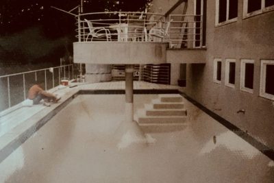 Empty swimming pool with a person sitting on the edge and patio furniture on a raised platform above the pool.