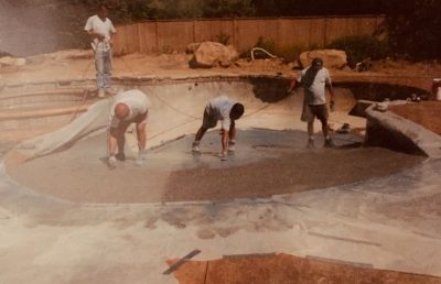 Four workers spread concrete at the bottom of an empty in-ground swimming pool under construction.