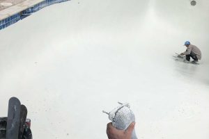 Two workers resurface an empty swimming pool with white material; tools and supplies are visible in the foreground.