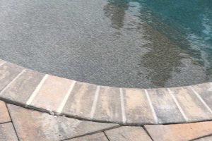 Curved edge of a swimming pool bordered by light brown concrete pavers; water is calm and clear.