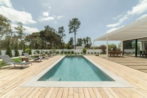 Rectangular swimming pool with wooden deck, lounge chairs, and patio dining area beside a modern house on a clear day.