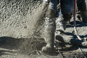 Worker spraying wet concrete onto a surface at a construction site, with a hose and concrete pile in the background.