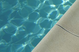 Close-up of a swimming pool with clear blue water next to a textured concrete pool edge in sunlight.